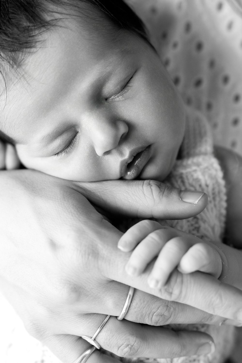 Black and white photo of a sleeping newborn baby cradled in a parent's hands wearing rings.