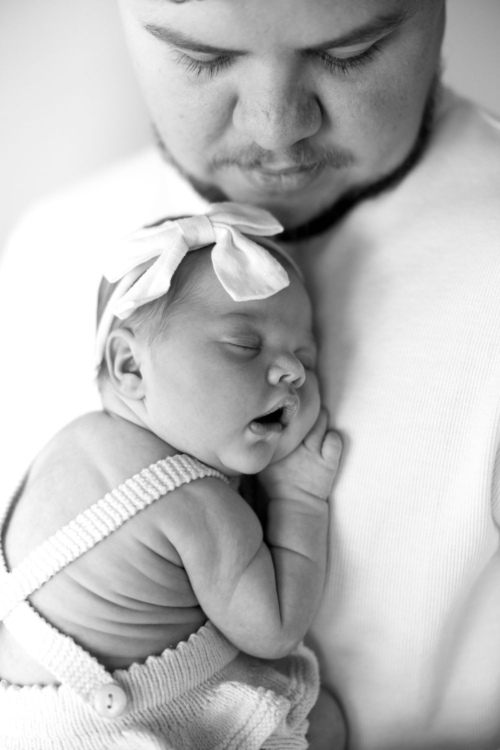 Black and white photo of a father tenderly holding his sleeping newborn baby girl wearing a bow headband.