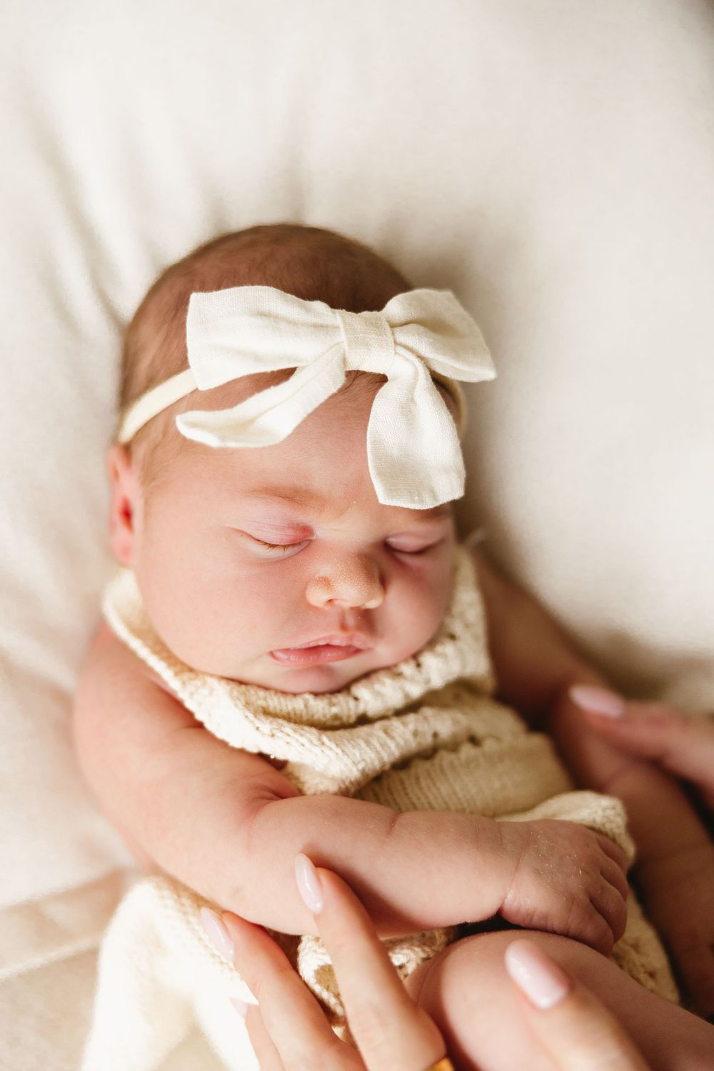 Sleeping newborn baby girl wearing a cream knit romper and white bow headband, cradled in mother's hands.