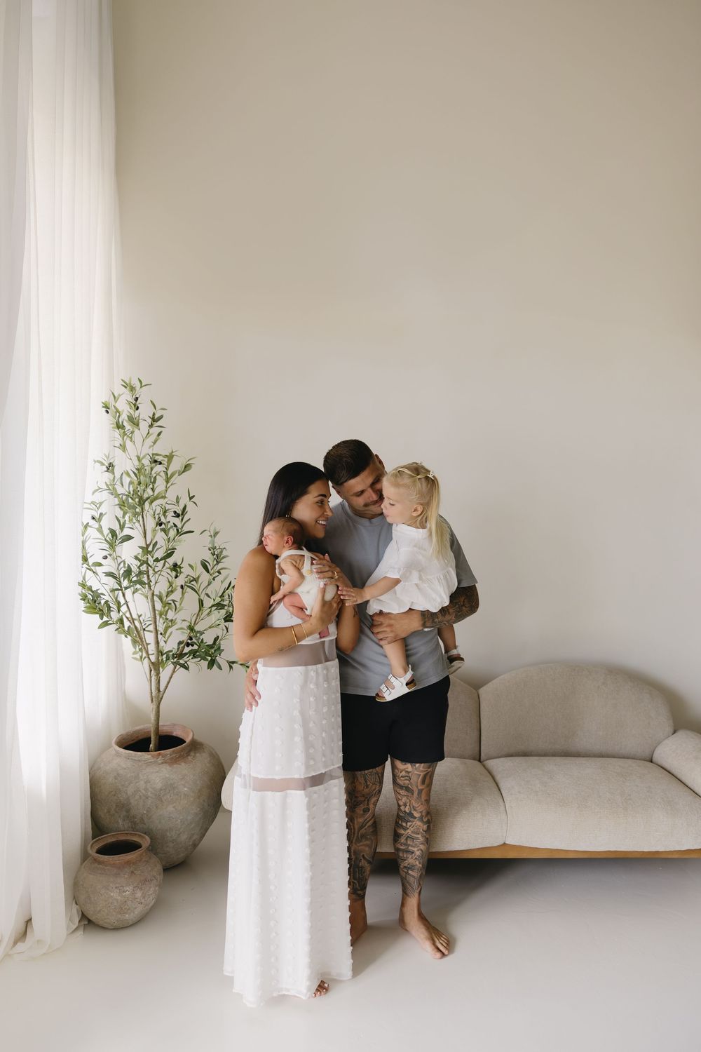 A minimalist white room features tender family moments captured in a series of connected photos with a potted olive tree.