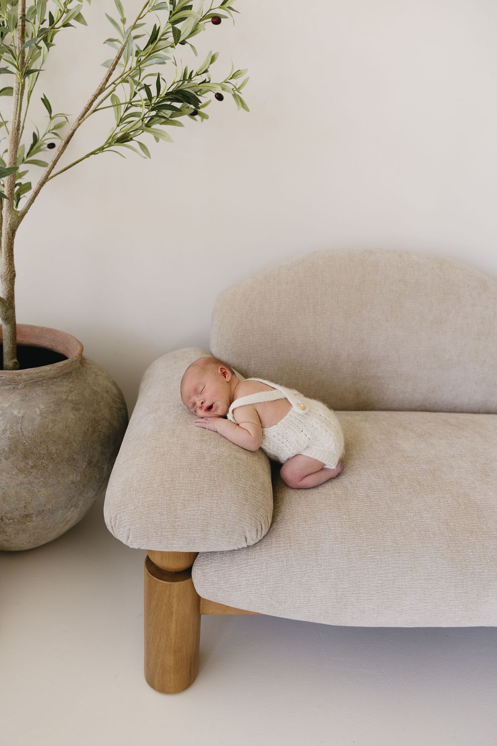 A sleeping newborn in white clothing rests peacefully on a beige couch next to a potted plant in a minimalist setting.