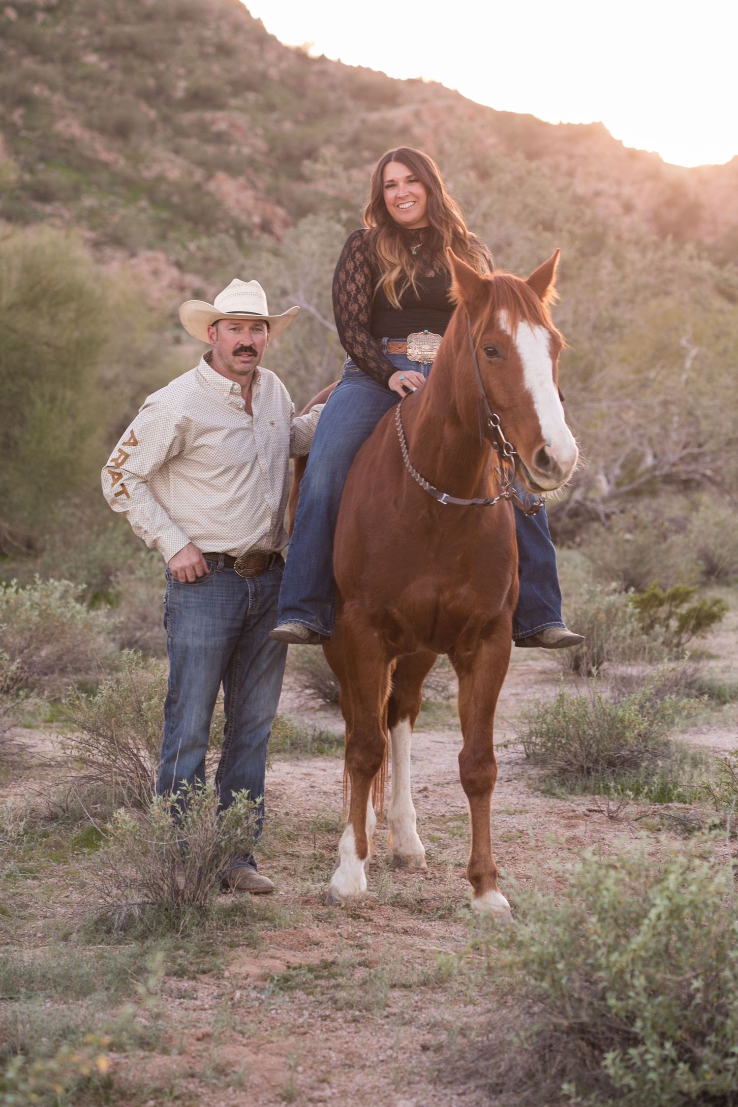 Man in cowboy hat stands beside woman riding chestnut horse in desert at sunset.