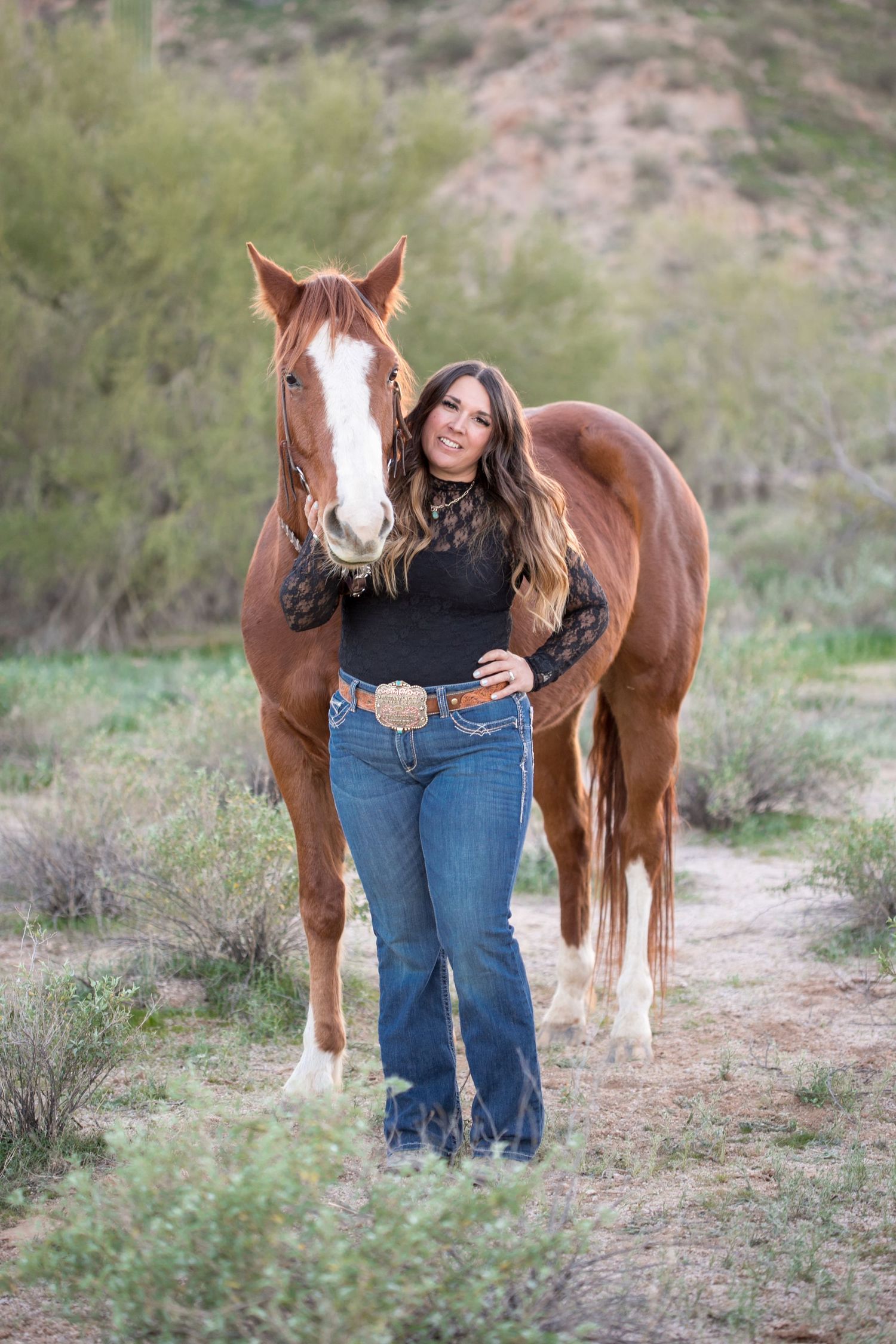 Woman in black lace top stands with chestnut horse on desert trail.