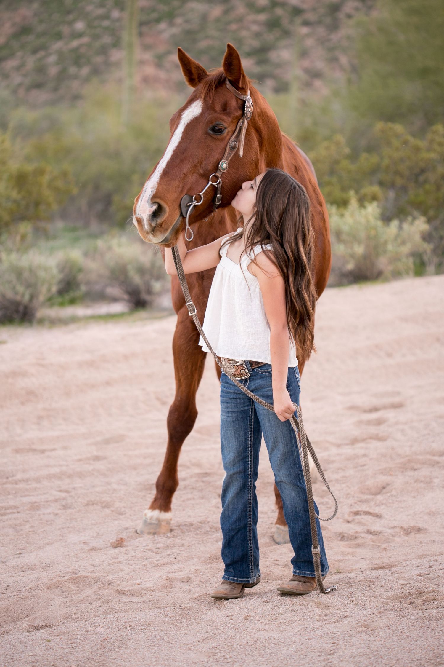 Young girl in white top kisses chestnut horse on sandy desert path.