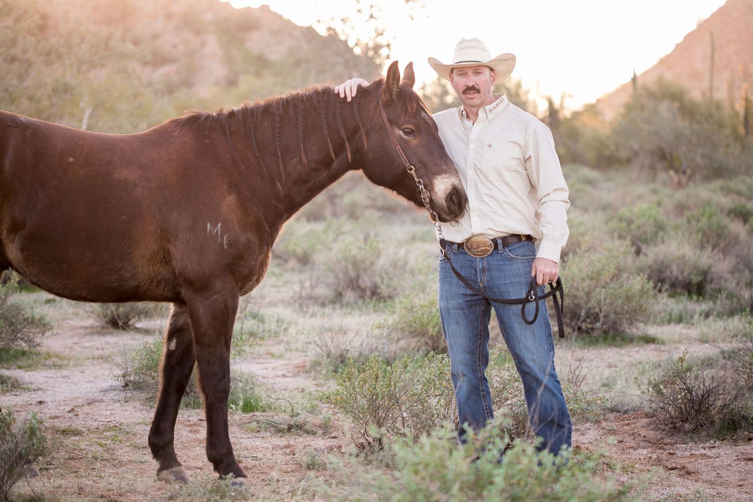 Cowboy in white hat and jeans stands beside a dark brown horse in a desert landscape at golden hour.