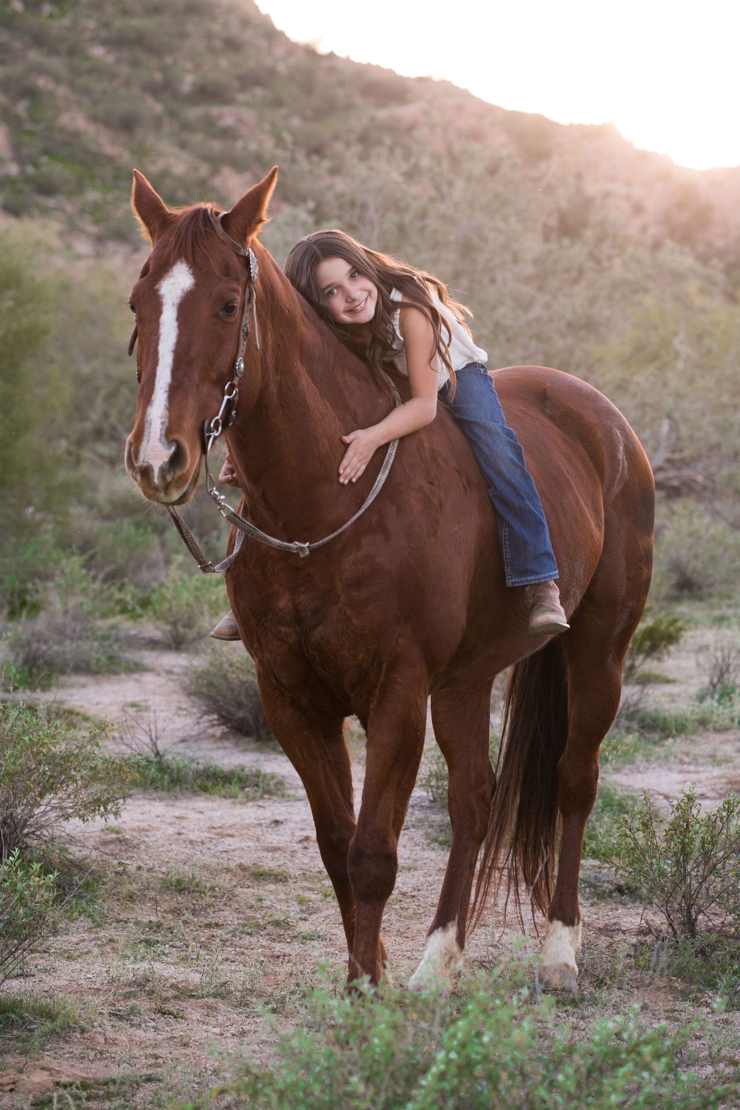 Young girl leans forward hugging chestnut horse in desert hillside setting.
