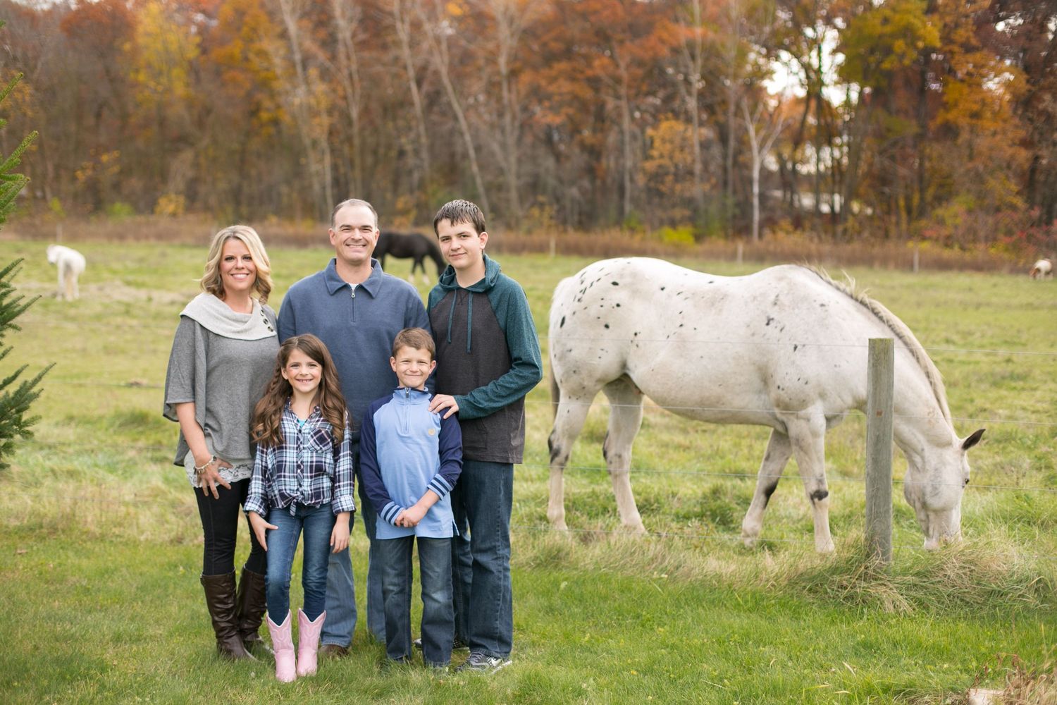 Family of five posing outdoors in a field with a white spotted horse grazing nearby in fall.