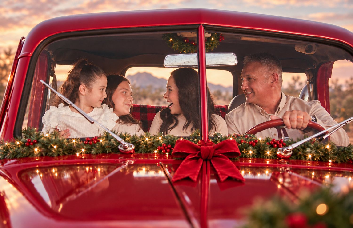 Family of four enjoying a festive Christmas car ride in a decorated red vintage truck with holiday lights and a red bow.