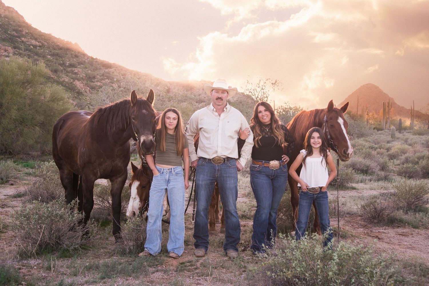 Family of five poses with two dark horses in desert landscape at golden hour.