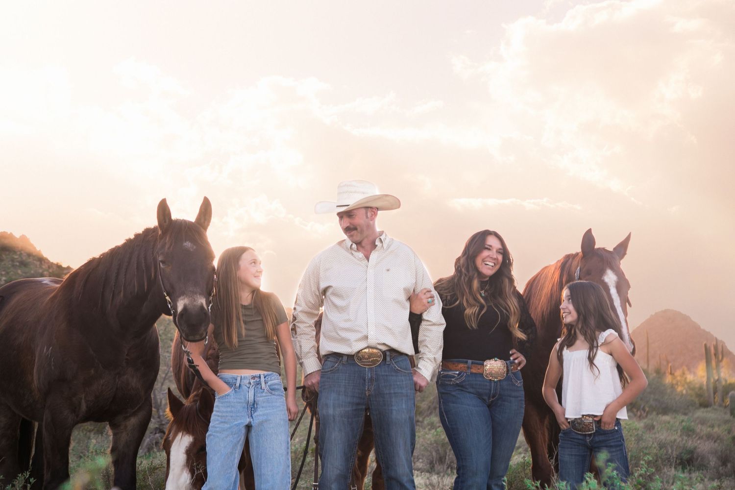 Man in cowboy hat walks with family and horses in desert at golden hour.