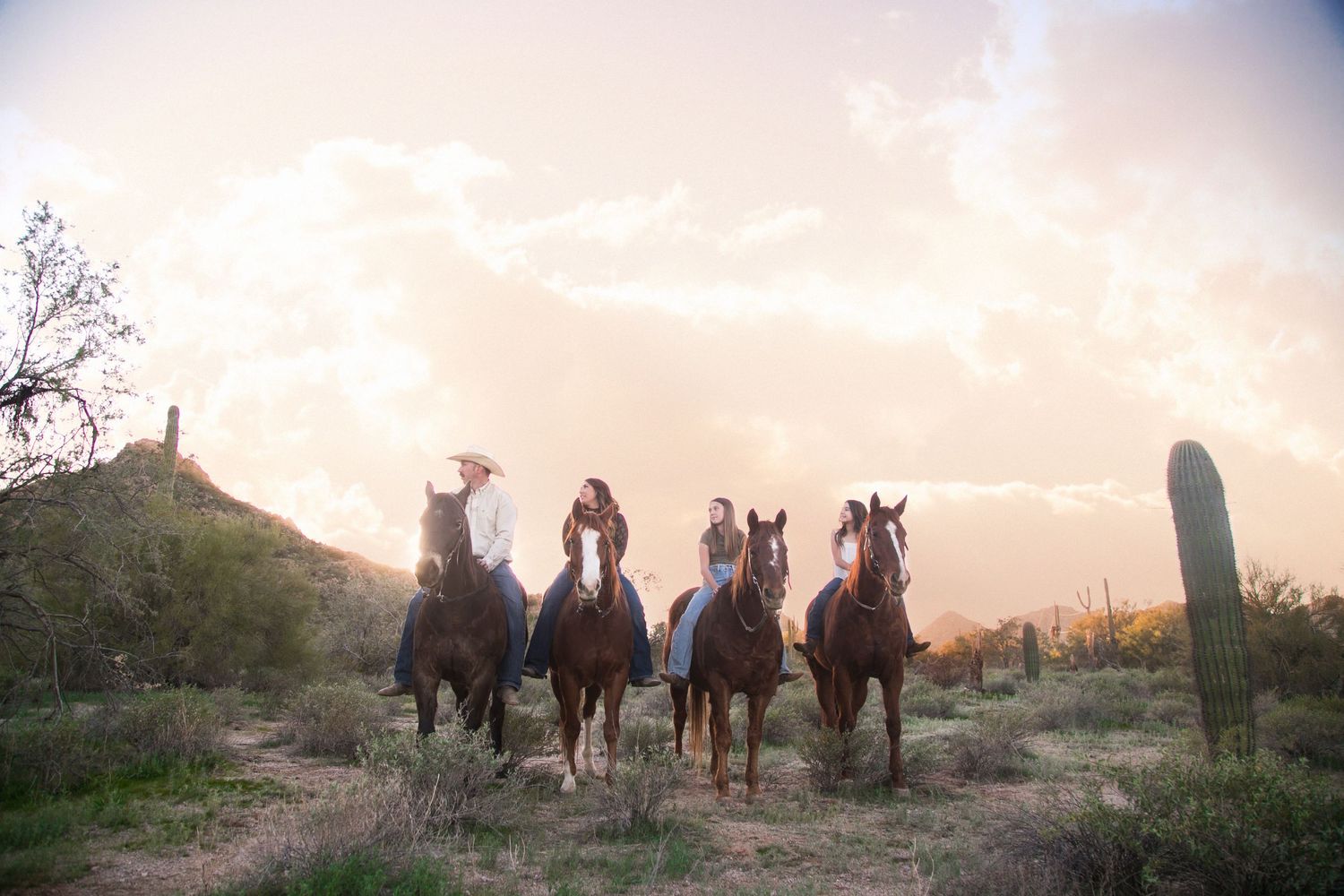 Family of four rides horses through desert landscape with saguaro cactus at sunset.