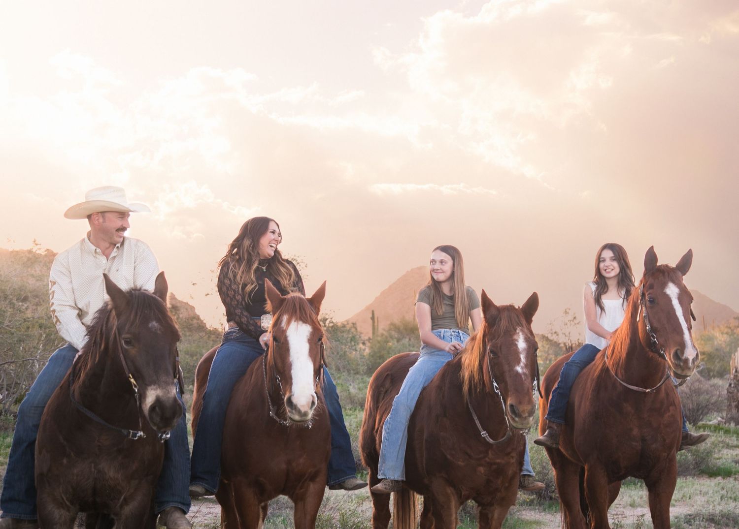 Family of four on horseback laughing together in desert landscape at sunset.