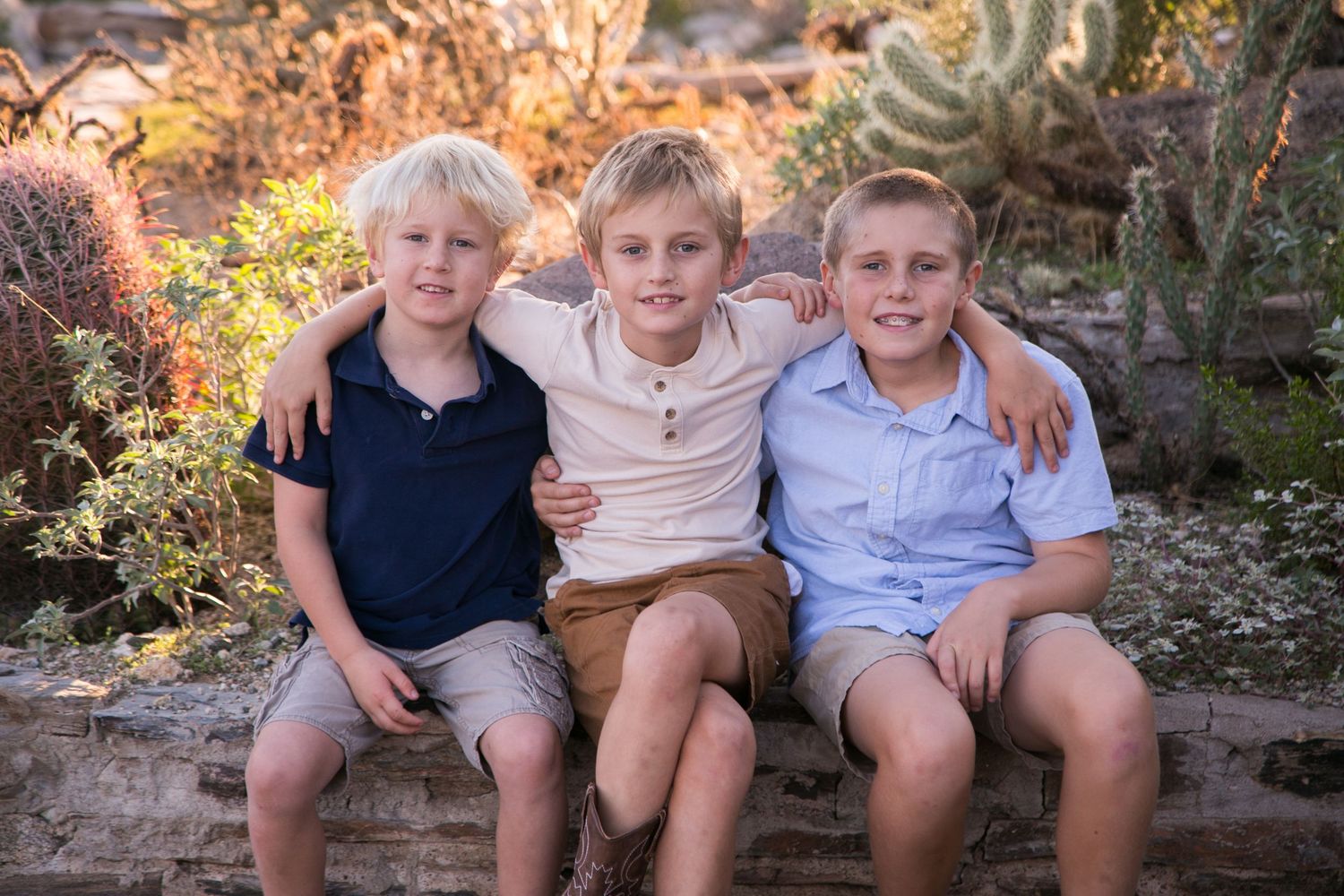 Three blond boys sitting together smiling in desert garden setting.