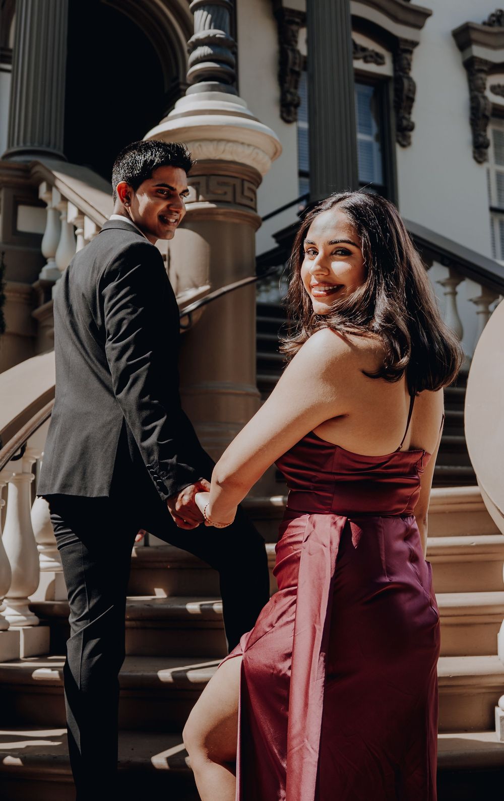 A couple in formal attire hold hands while ascending ornate stone steps outside a grand historic building entrance.