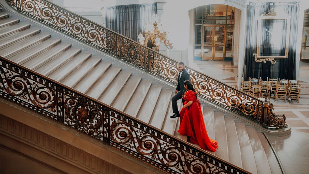 Woman in red dress poses on grand marble staircase with ornate iron railings in luxurious building.