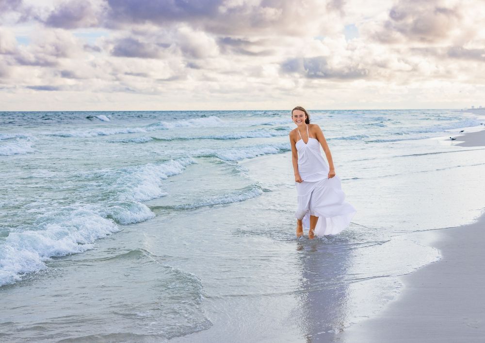 A woman in a white dress walks along the shoreline as gentle waves roll onto a sandy beach at sunset.