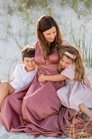 A coastal family portrait shows three people in light summer dresses sitting together on white sand among beach grass.