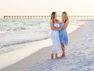 Two women in white and blue dresses embrace on a sunny beach with a pier in the background.