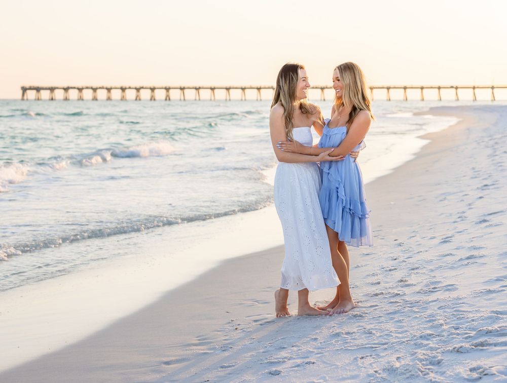 Two women in white and blue dresses embrace on a sunny beach with a pier in the background.