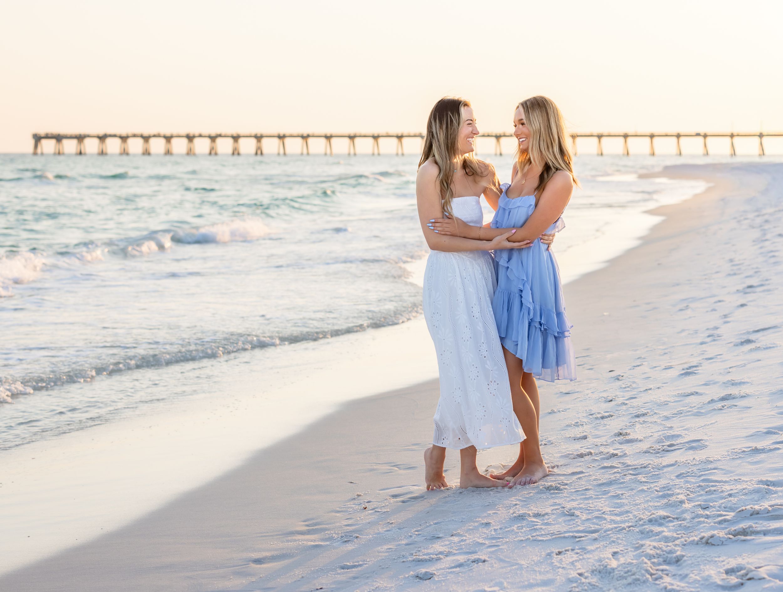 Two women in white and blue dresses embrace on a sunny beach with a pier in the background.