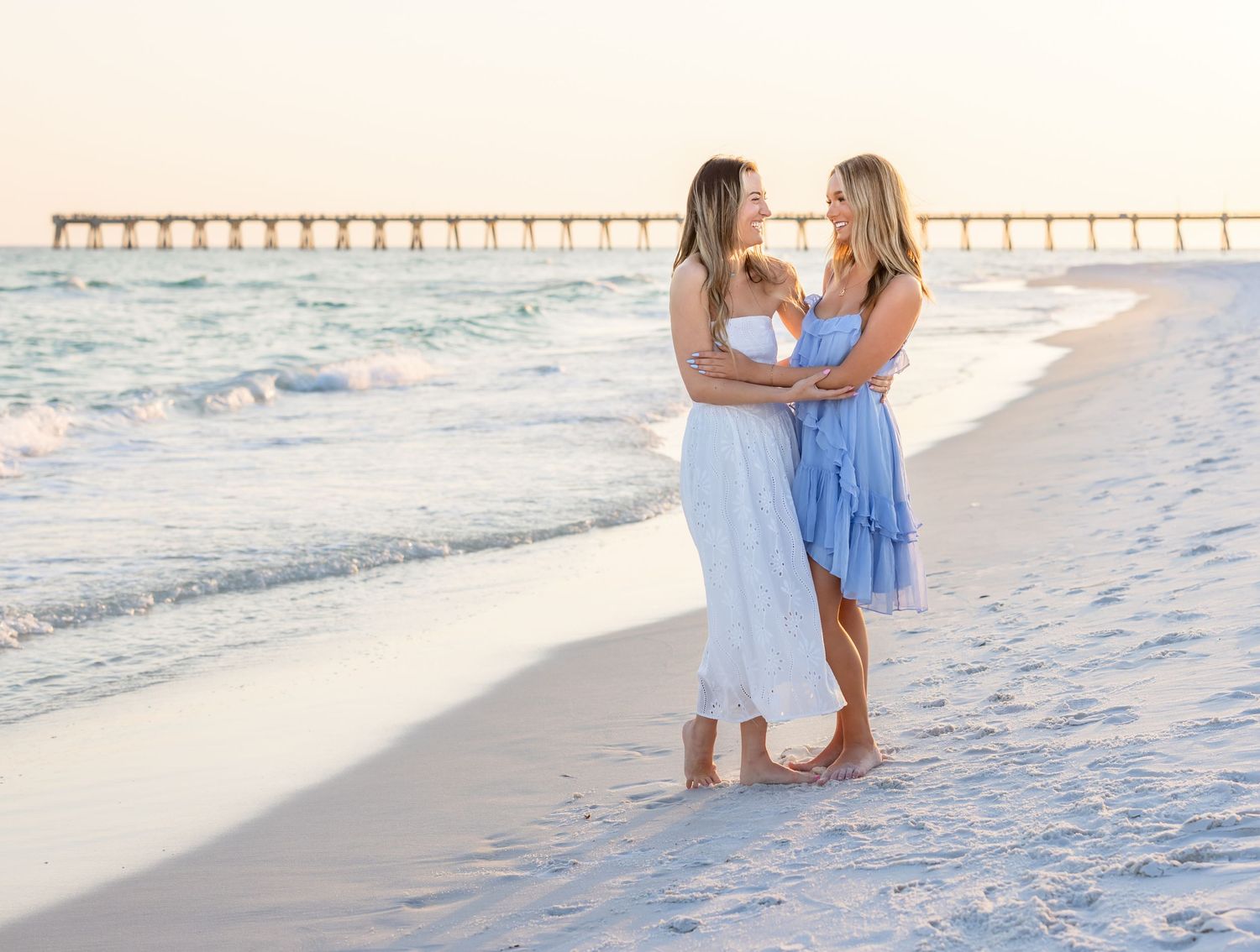 Two women in white and blue dresses embrace on a sunny beach with a pier in the background.