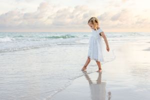 Small child in white dress twirling at the ocean's edge during golden hour.