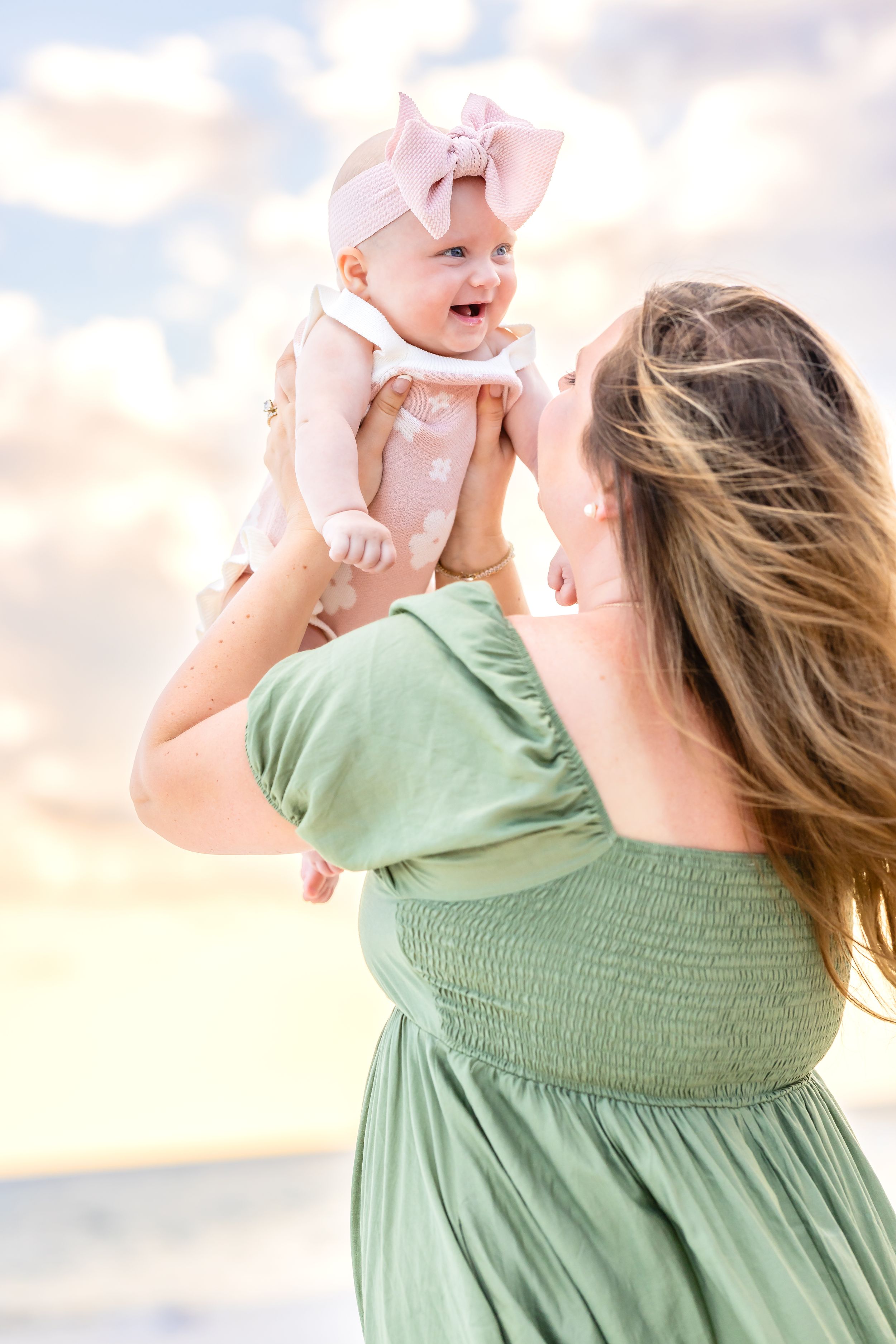 A tender moment captured at sunset as a woman in a sage green dress lifts a baby wearing pink against a dreamy sky.