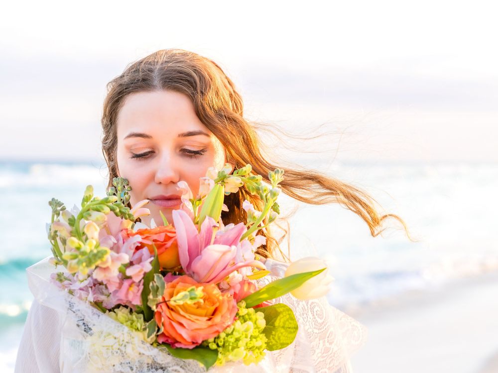Young bride smelling a colorful bouquet of pink and orange flowers on a breezy beach at sunset.