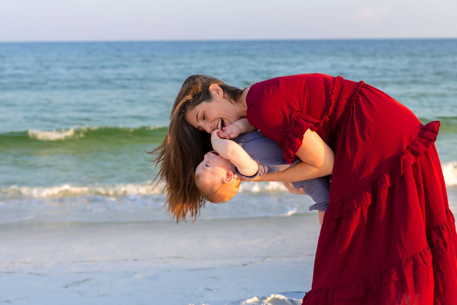 Woman in red dress joyfully holding a baby on a sunny beach with turquoise ocean waves in the background.