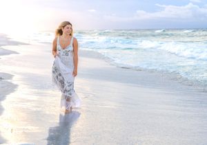 Woman in white floral dress walking barefoot on a sunny beach with gentle waves.