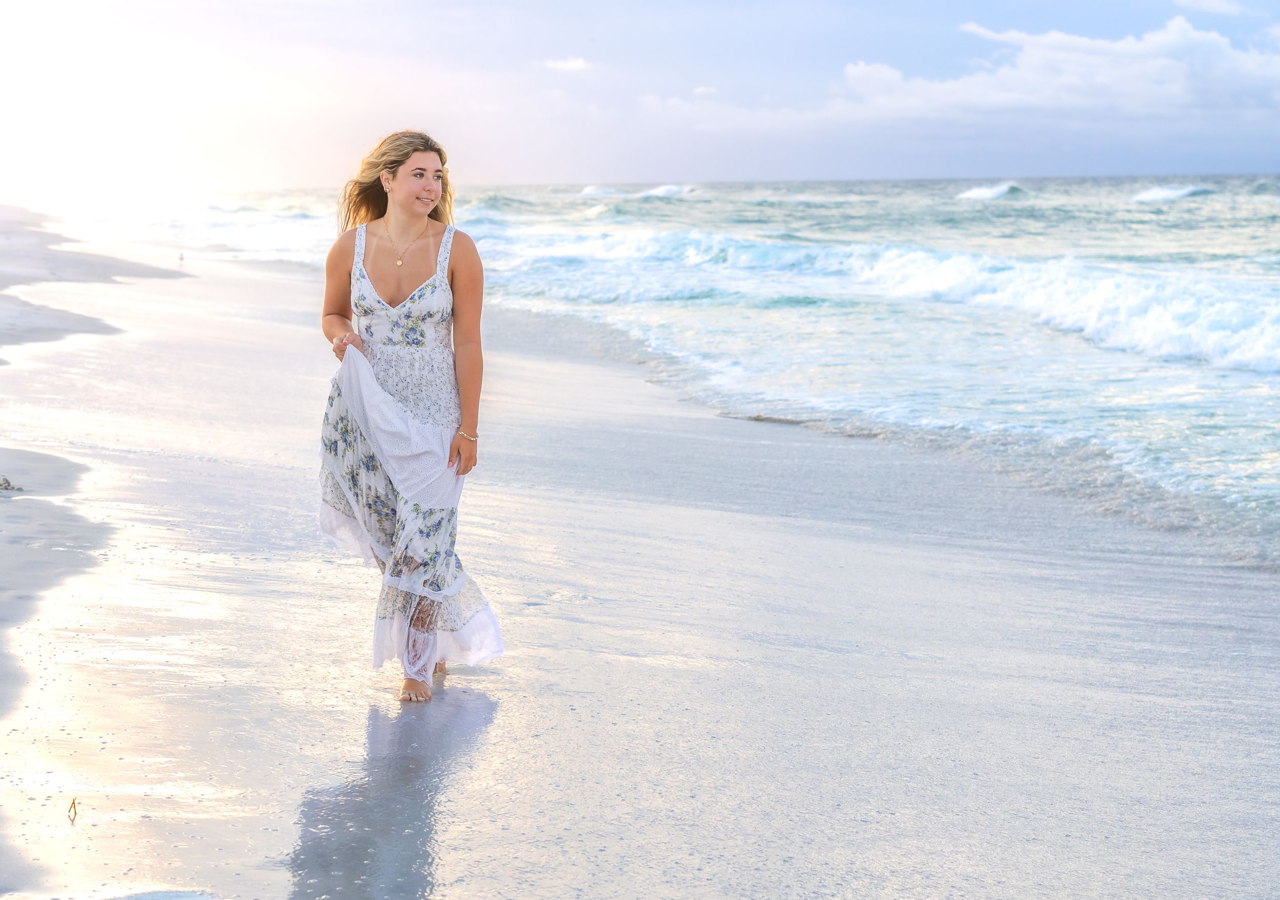 Woman in white floral dress walking barefoot on a sunny beach with gentle waves.