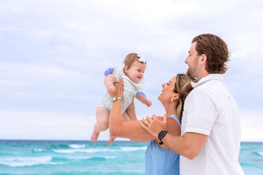 A happy family with a man and woman lifting a smiling baby on a sunny beach with turquoise ocean waves.