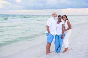 Three people in coordinating blue and white outfits pose together on a sunny beach beside turquoise ocean waves.
