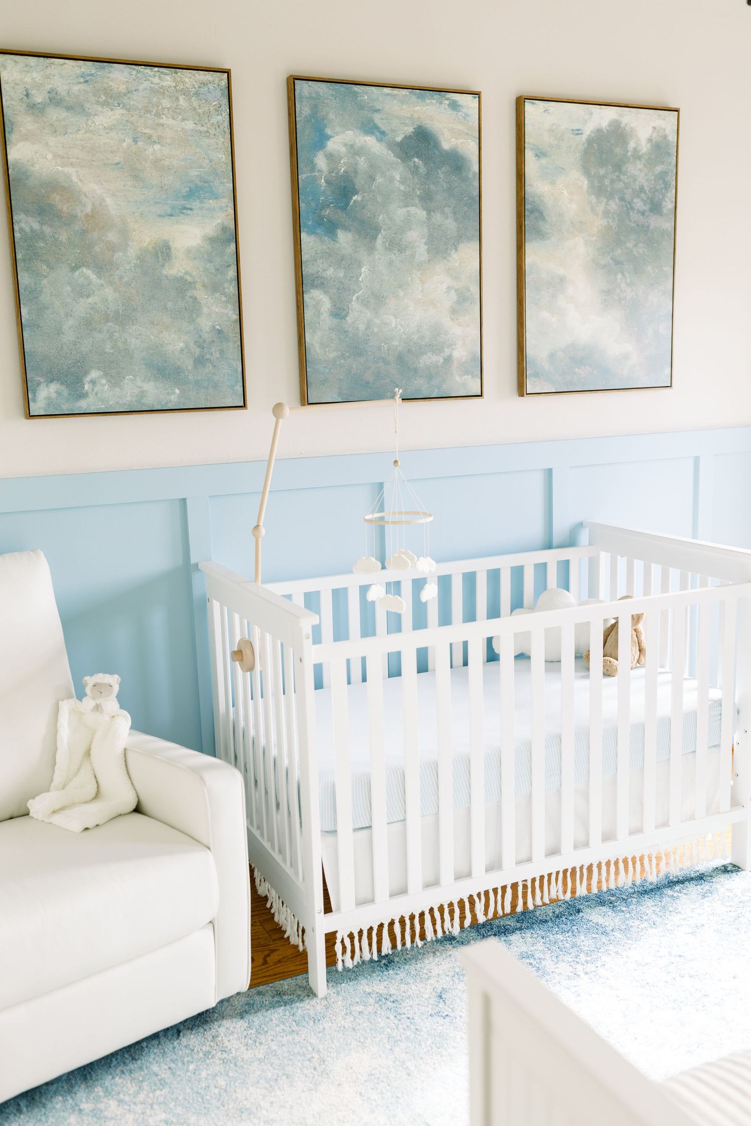Modern nursery with cloud artwork, white crib, and blue wainscoting creates a serene atmosphere.