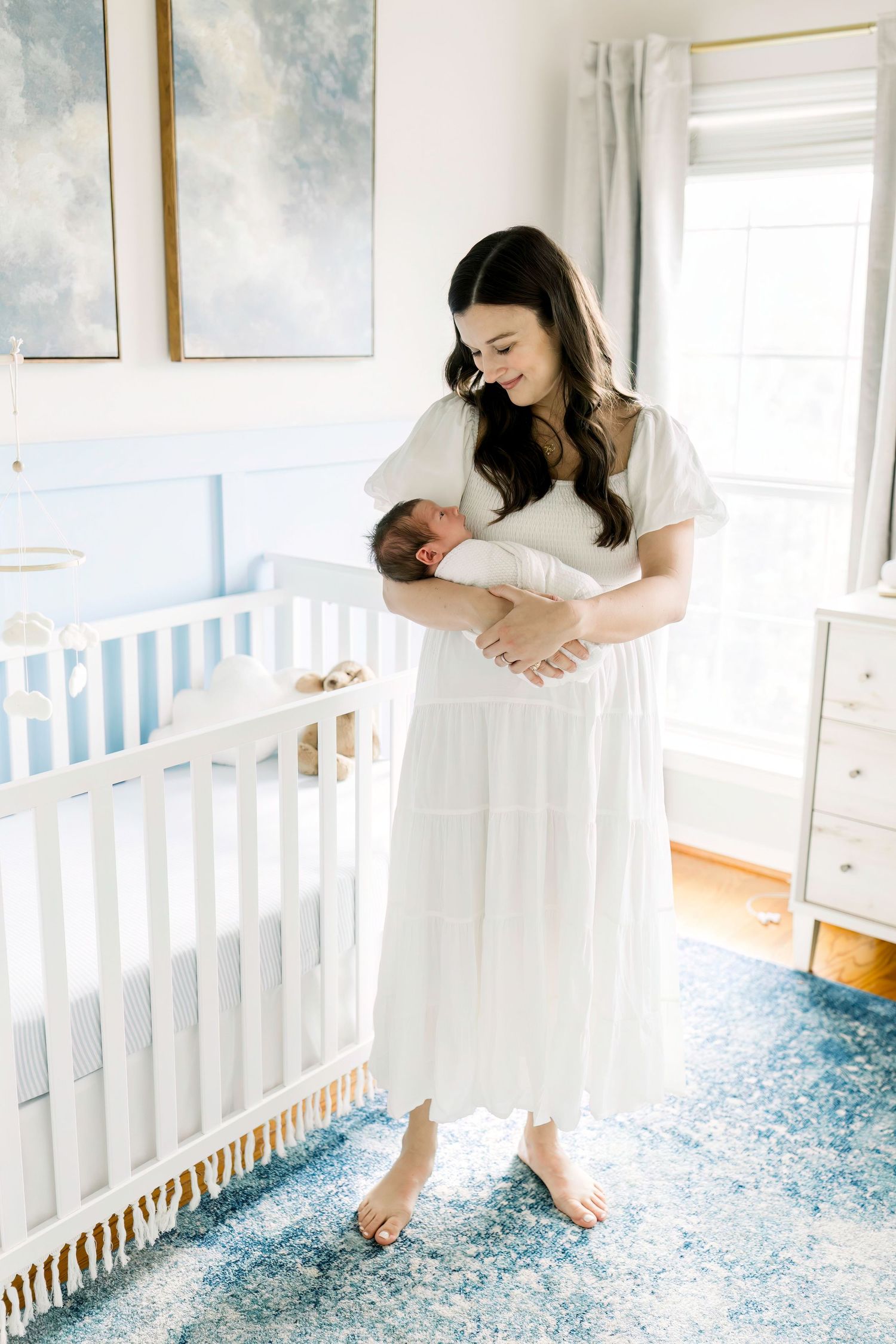Tender moment in a nursery with cloud artwork as mother in white dress holds sleeping newborn.