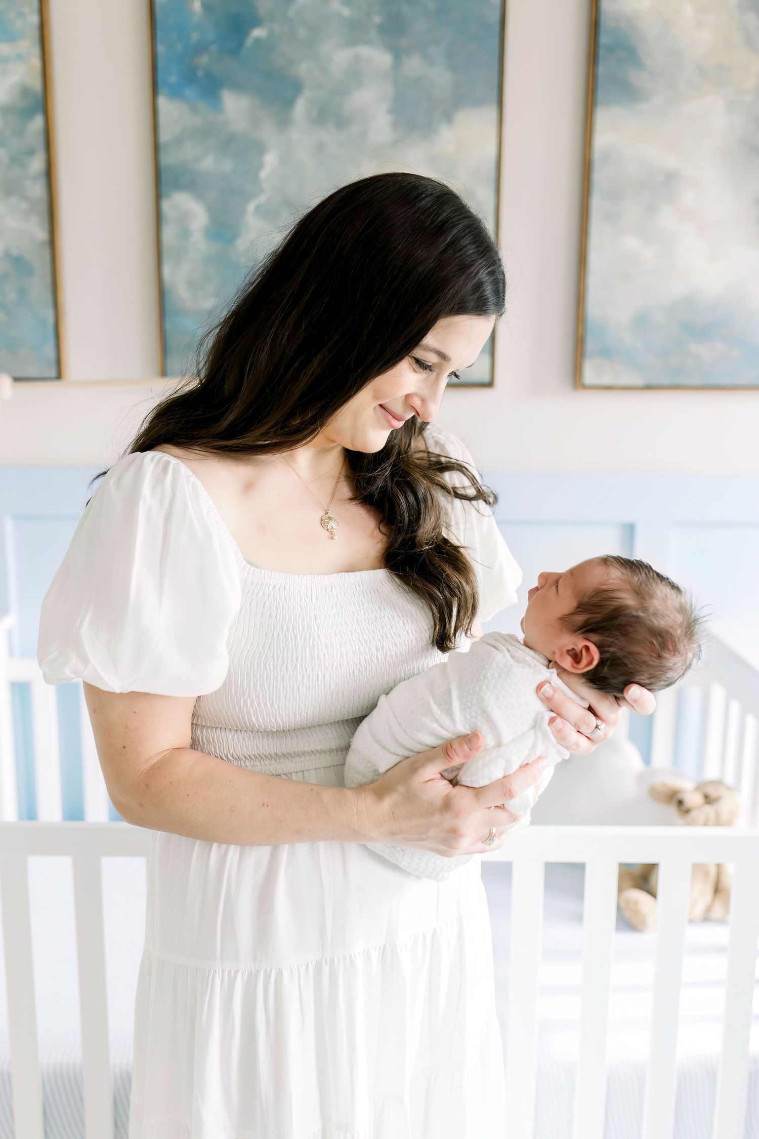 Intimate nursery moment with mother in white dress holding newborn by cloud-themed wall art.