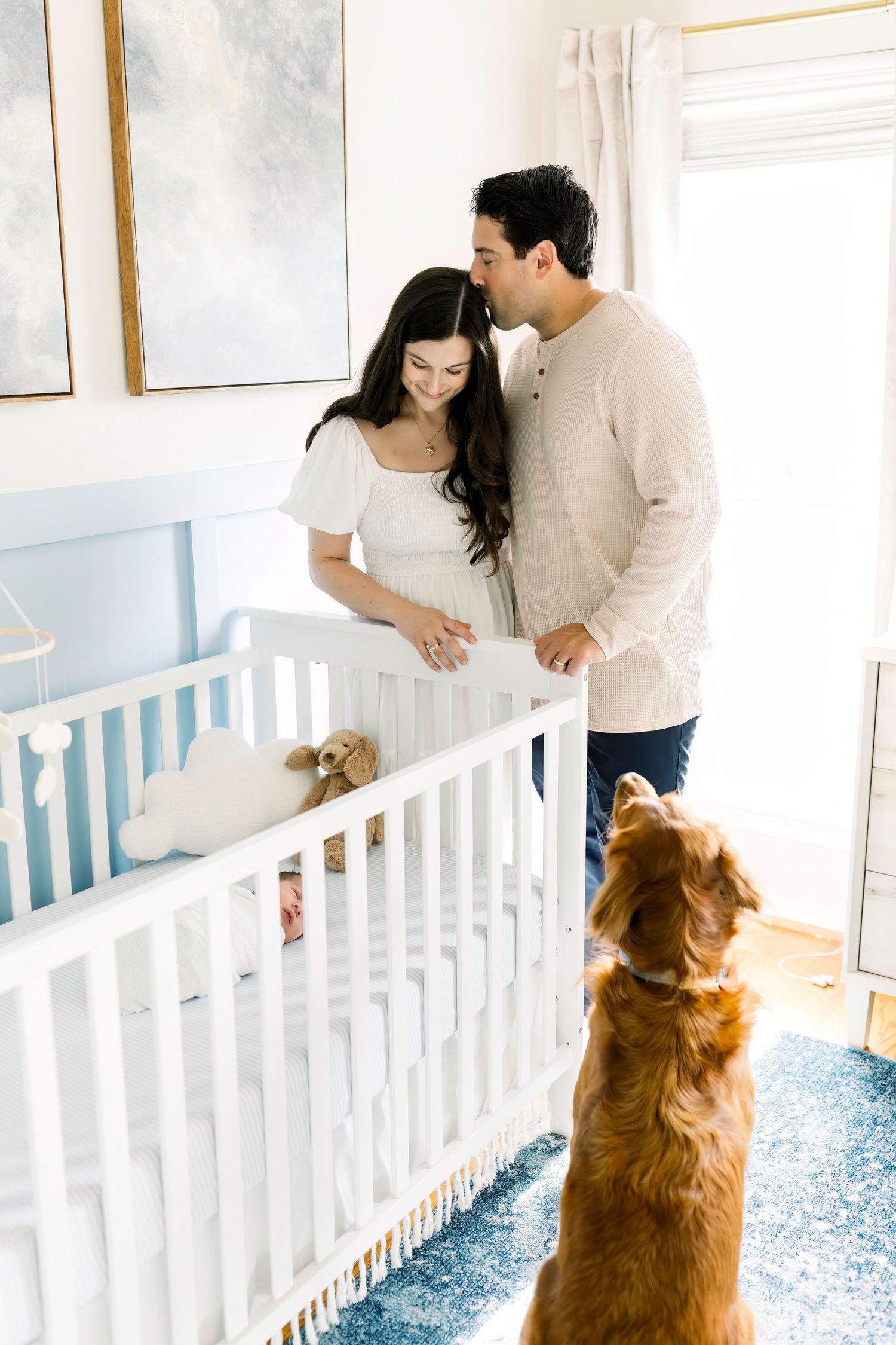 Nursery scene with couple admiring their baby while golden retriever watches from below.