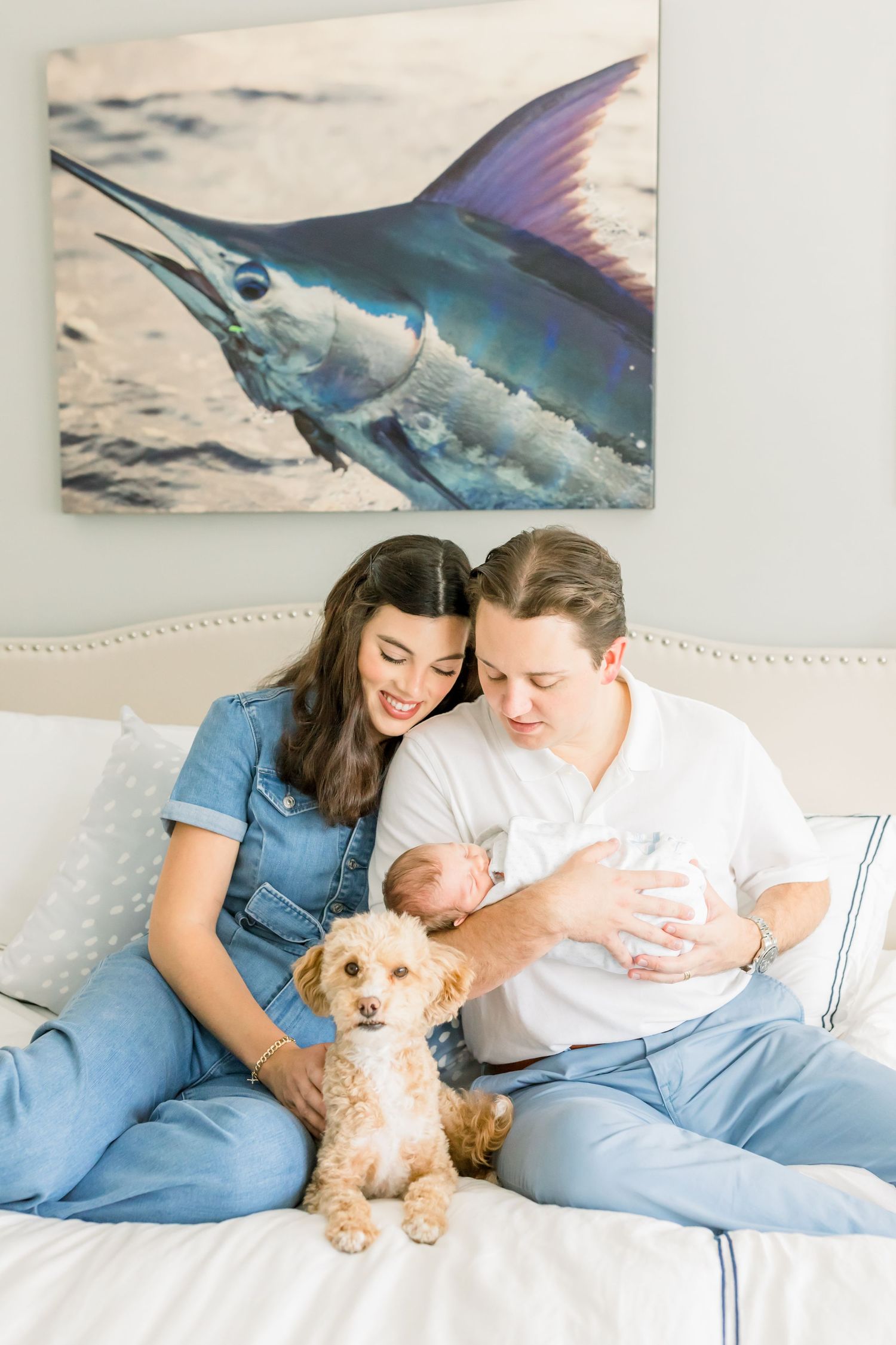 A couple sitting on a bed with their small dog and baby beneath a marlin artwork.
