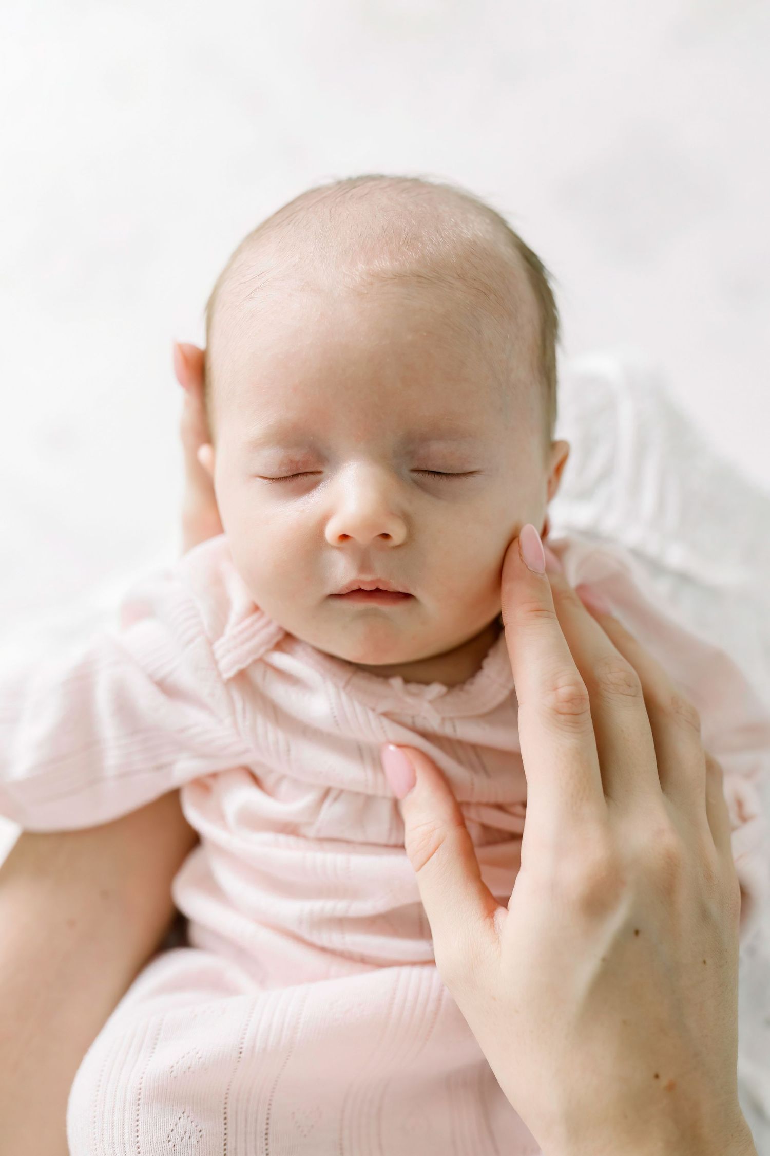 Close-up of sleeping newborn baby in pink outfit with gentle hands cradling their face.