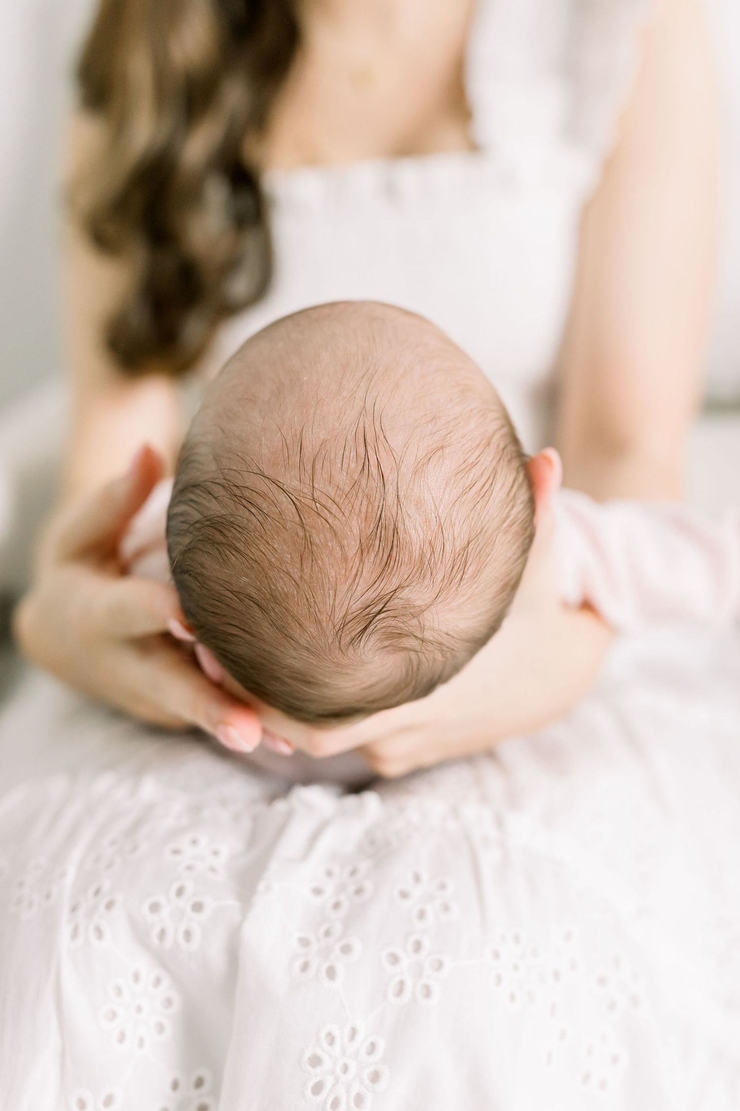 Top view of baby's head resting on white eyelet fabric with caring hands supporting.