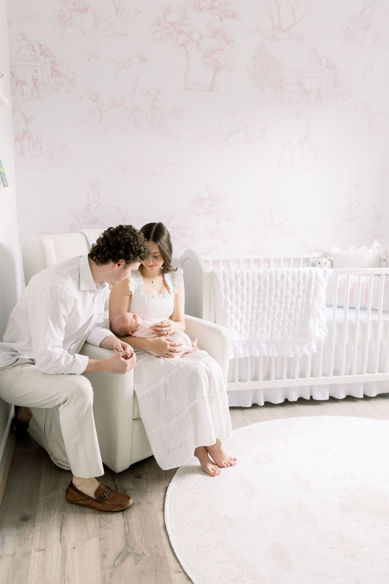 A tender moment in a white nursery room with a couple sitting on a plush armchair near a white crib.