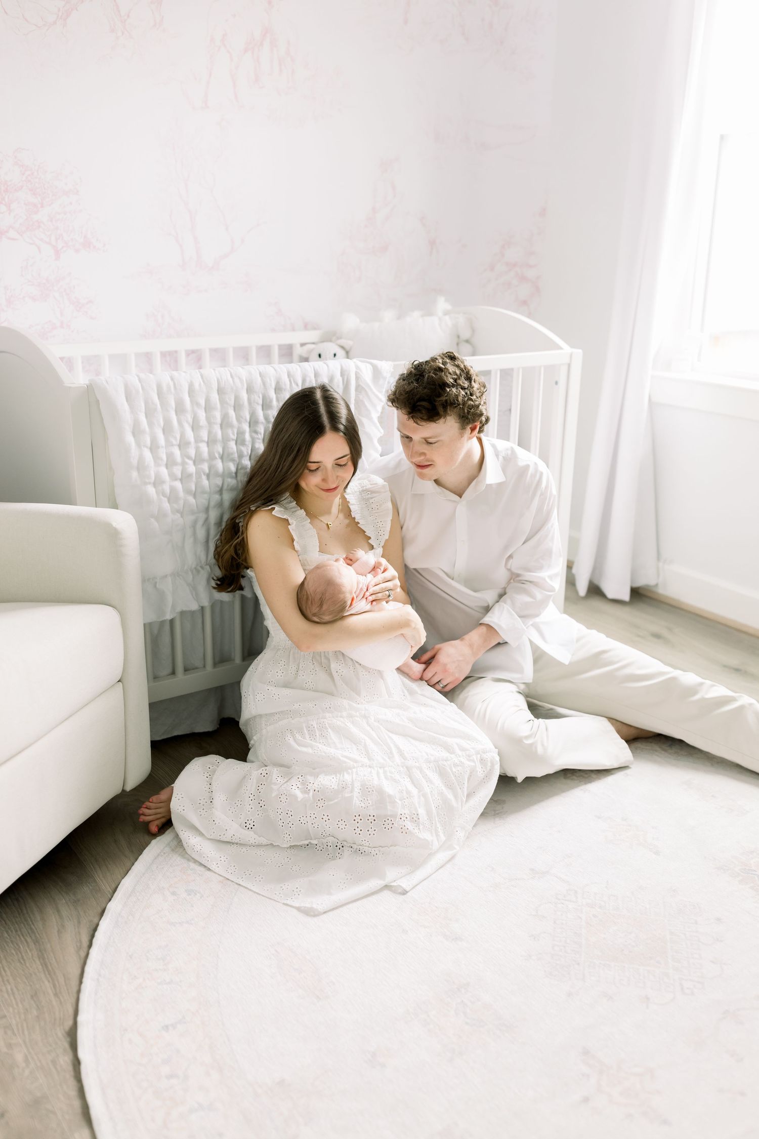 A couple shares an intimate moment on a white round rug beside a baby crib in a bright nursery.