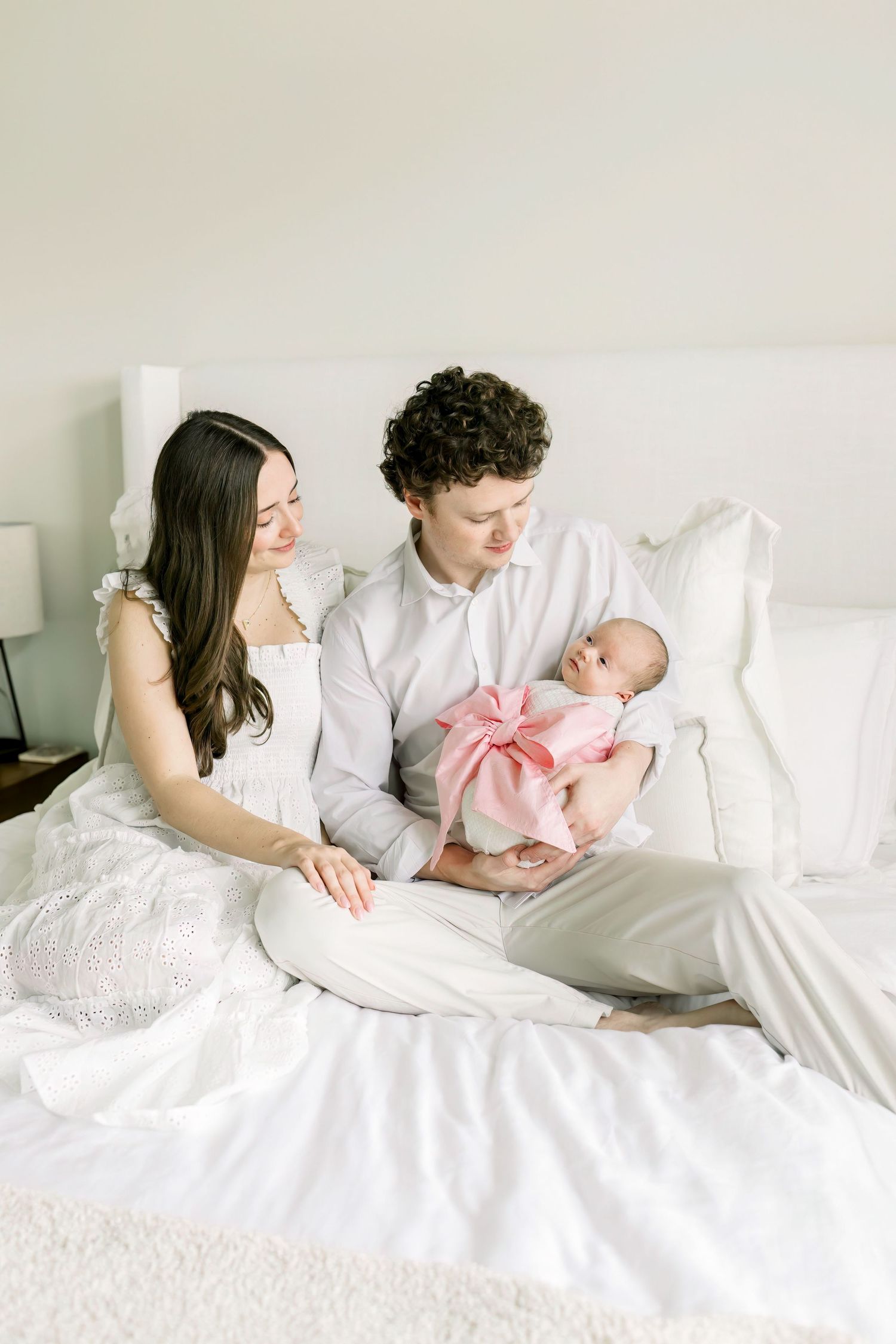 Family lounges together on white bedding with newborn baby wearing pink bow wrap.