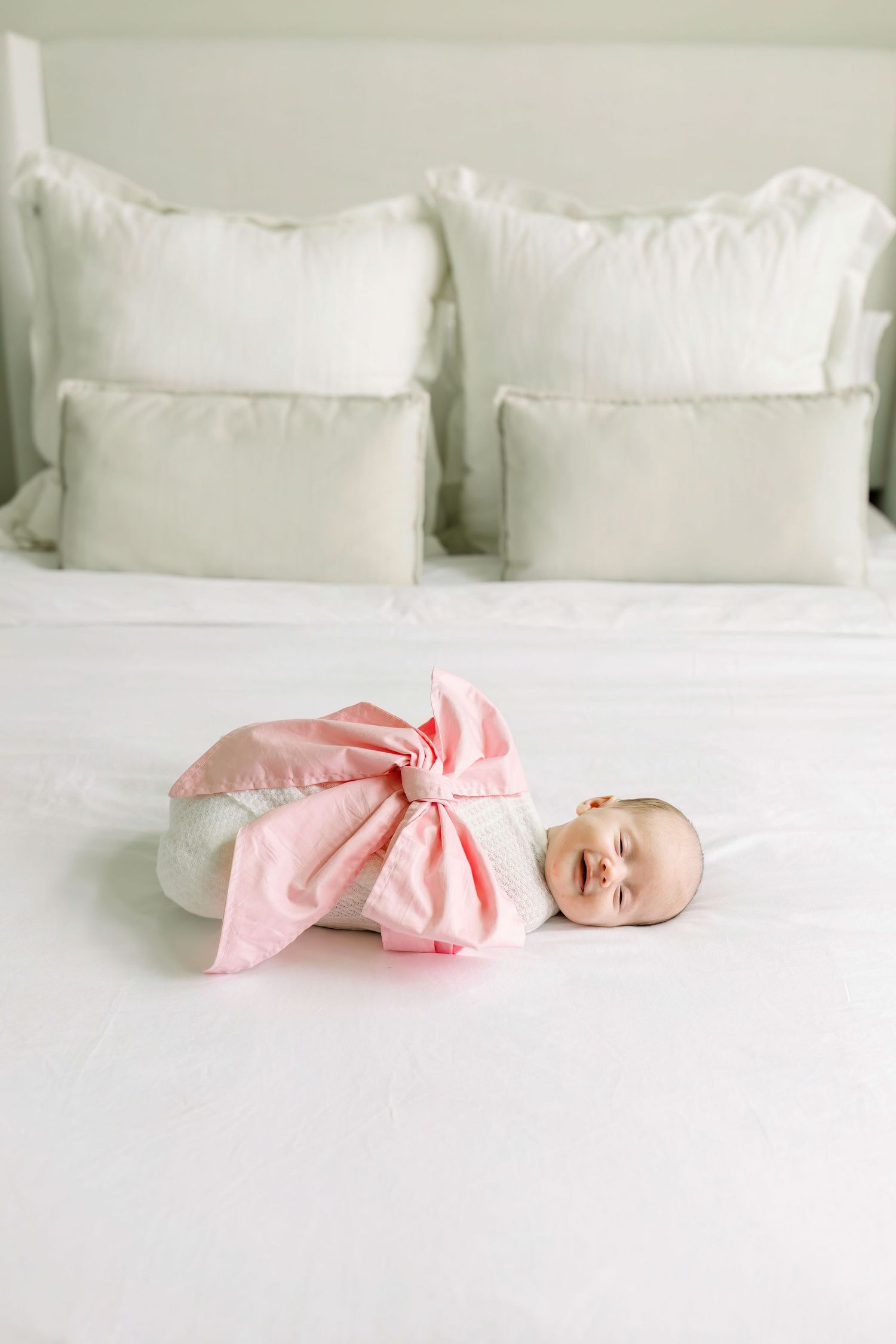 Newborn baby in pink bow swaddle lying on white bedding with pillows.