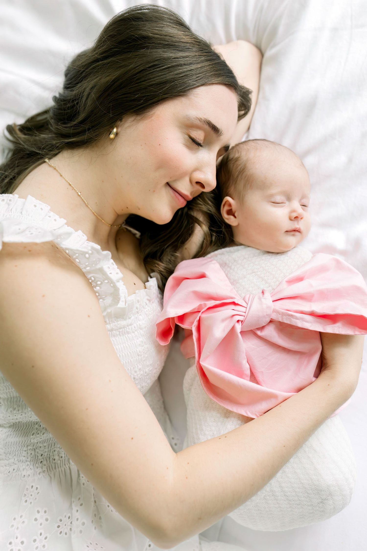 Tender moment between mother and baby lying on white bedding wearing matching white and pink outfits.