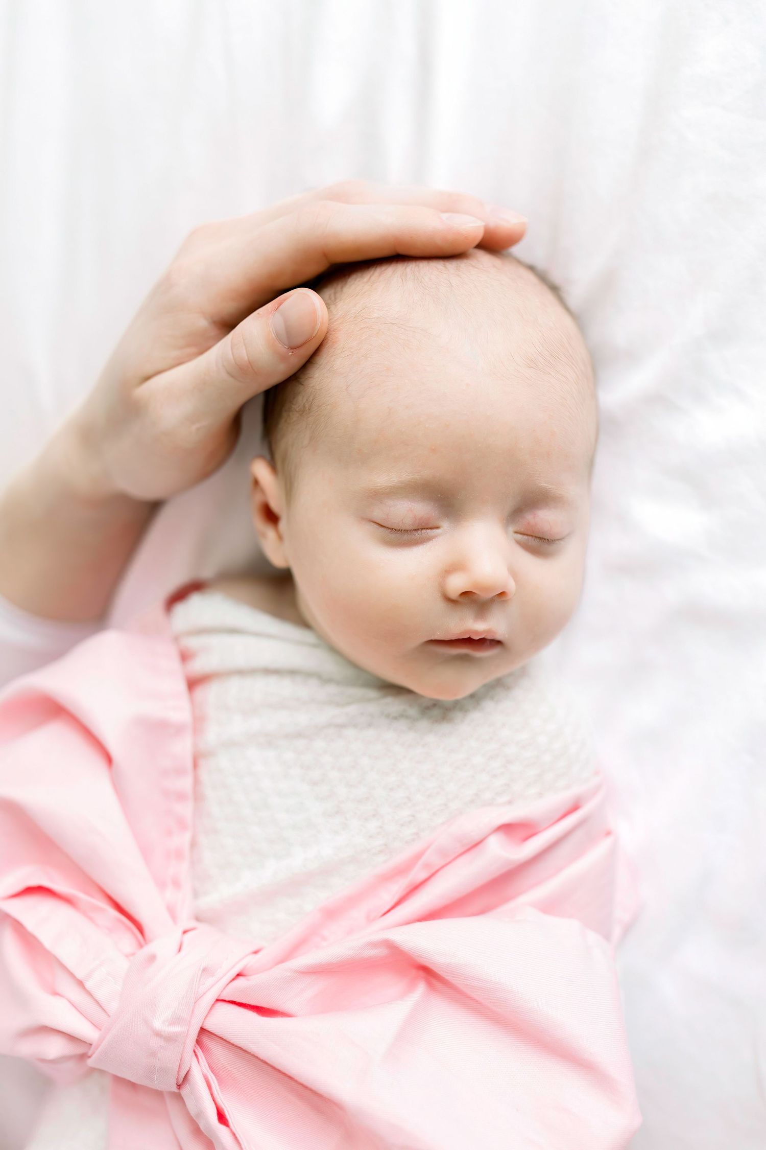 Close up of gentle hand caressing sleeping newborn in pink and white swaddle.