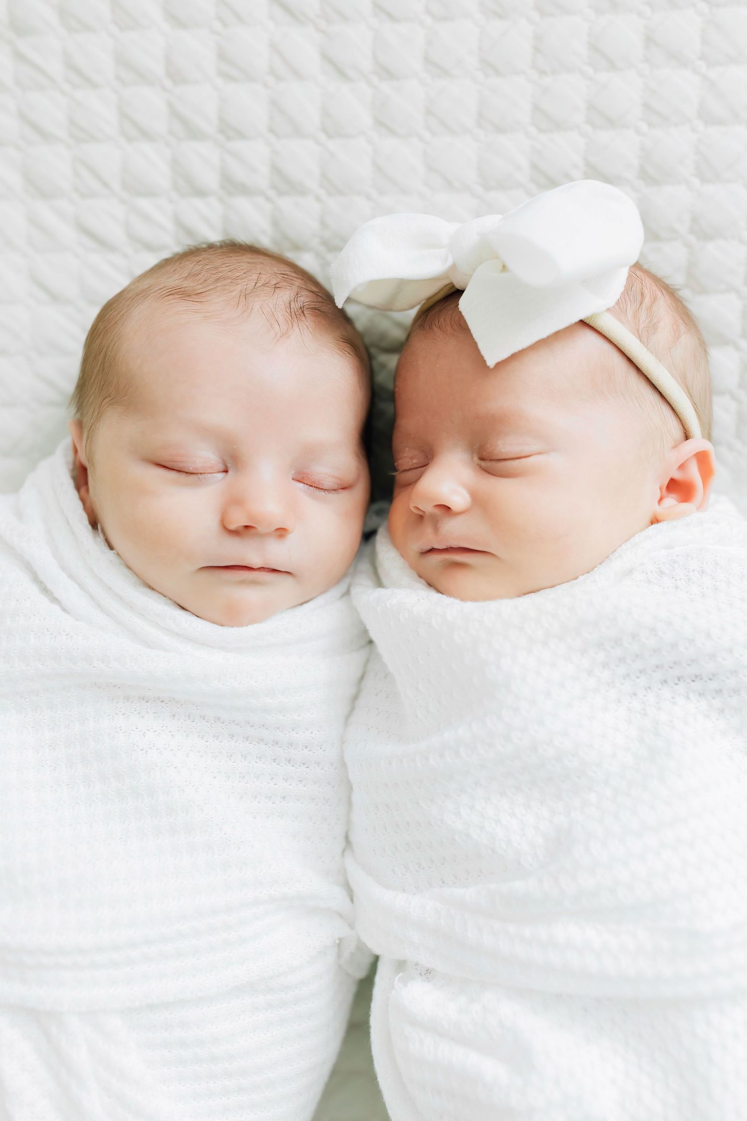 Two sleeping newborn babies in white swaddles lying side by side on white bedding.