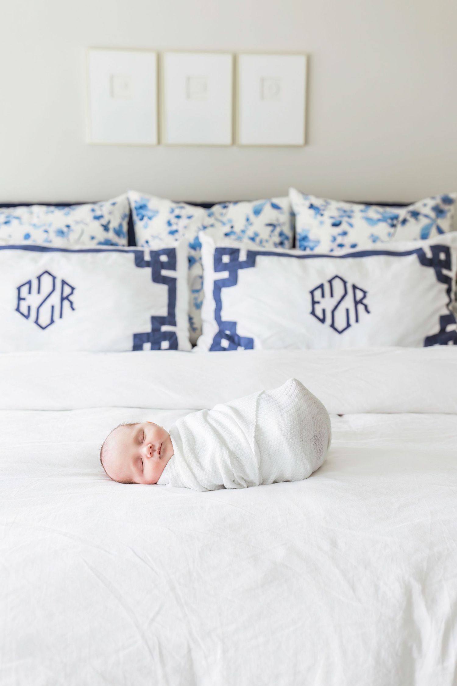 A sleeping newborn in white swaddle on bed with blue monogrammed pillows and white bedding.
