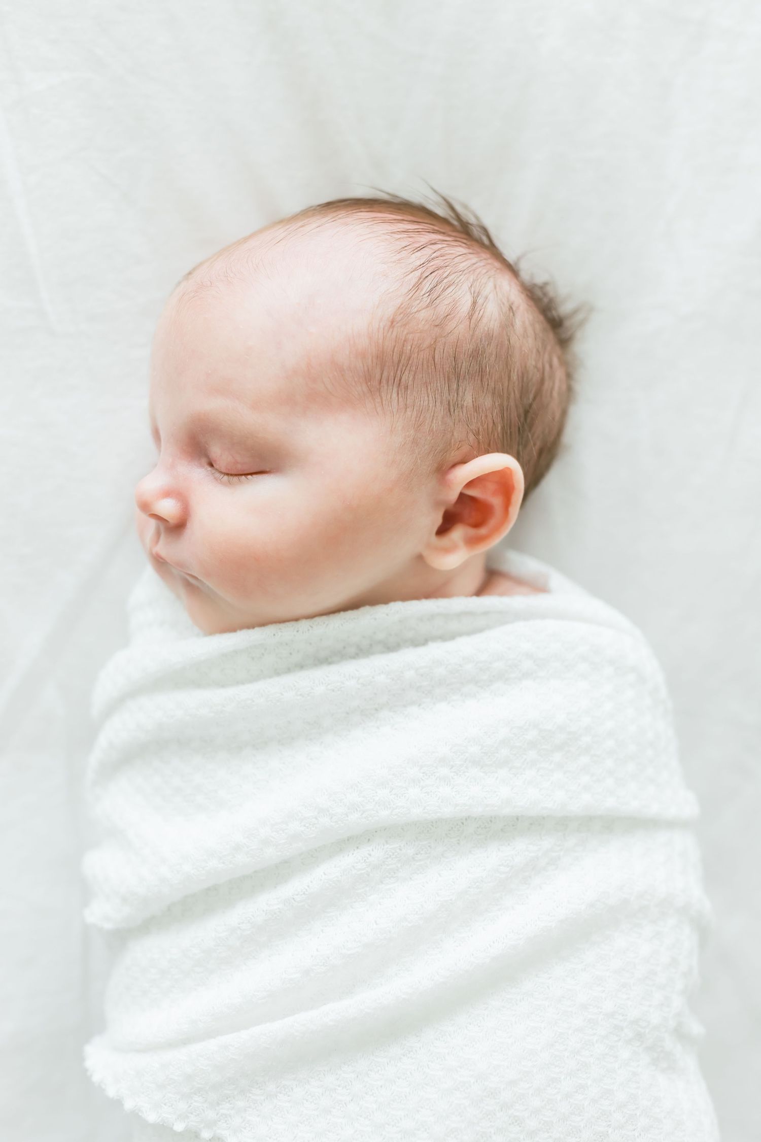 Close-up portrait of sleeping baby wrapped in white swaddle against white background.