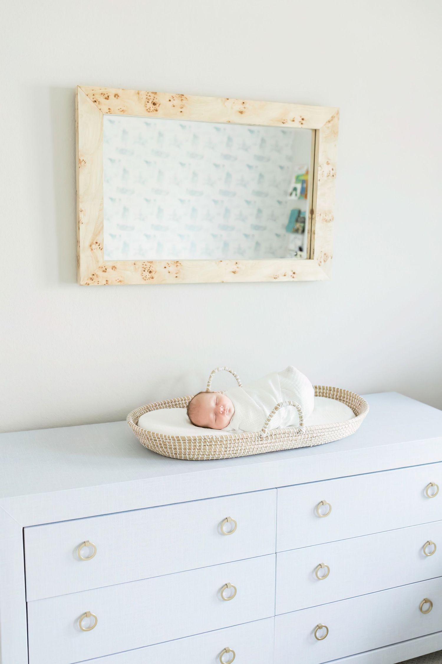 Newborn baby sleeping in white bassinet on white dresser beneath vintage mirror.